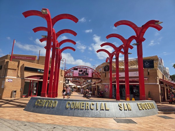 Entrada del Centro Comercial San Eugenio con altas estructuras rojas, tiendas y personas caminando en una jornada soleada.