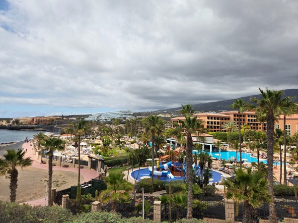 Vista elevada de la zona de Costa Adeje con hoteles, palmeras, piscinas y un cielo nublado sobre la costa.