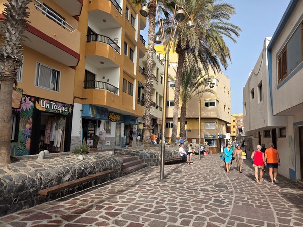 Calle peatonal de El Médano con palmeras, edificios de colores, pequeños comercios y personas paseando.
