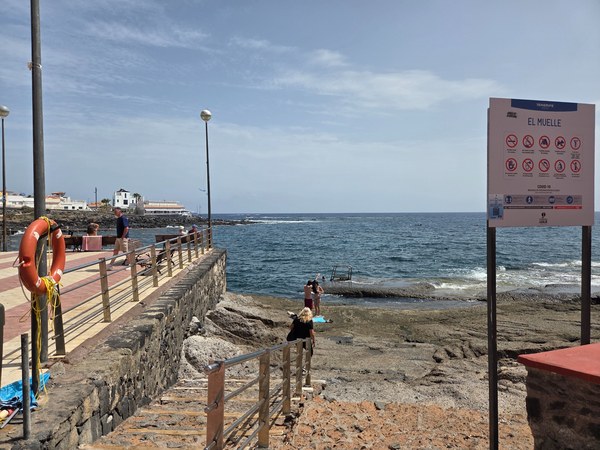 Zona de El Muelle en La Caleta con un cartel informativo, escaleras hacia el mar y personas junto a la orilla rocosa.