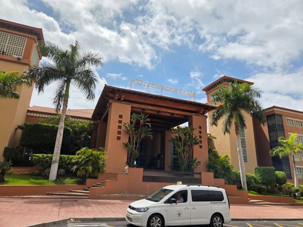 Fachada principal del H10 Costa Adeje Palace con palmeras, entrada porticada y cielo parcialmente despejado.