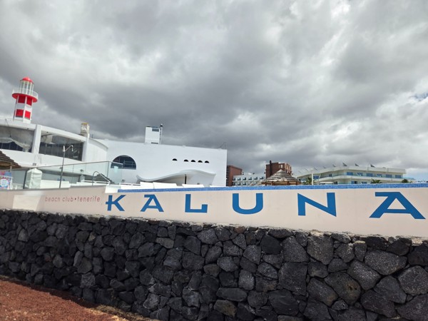 Muro exterior del Kaluna Beach Club Tenerife con letras azules, piedra volcánica y nubes grises sobre la costa.