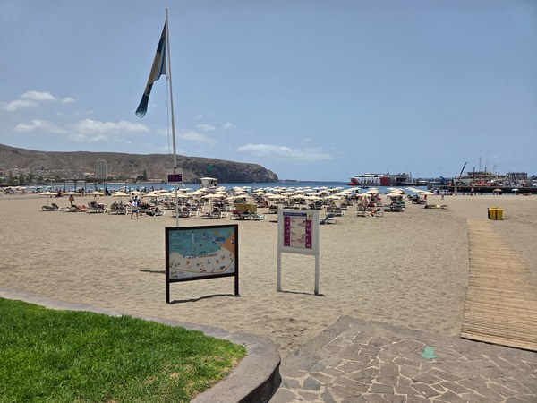 Playa de Los Cristianos Playa de Los Cristianos con arena clara, tumbonas, sombrillas y el puerto visible al fondo.