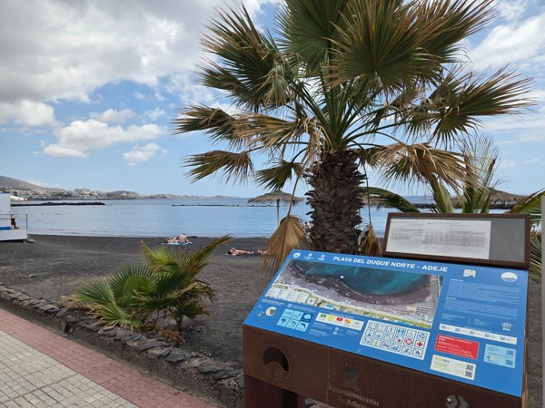 Panel informativo de Playa del Duque Norte en Adeje junto a la arena oscura, palmeras y el mar en calma.