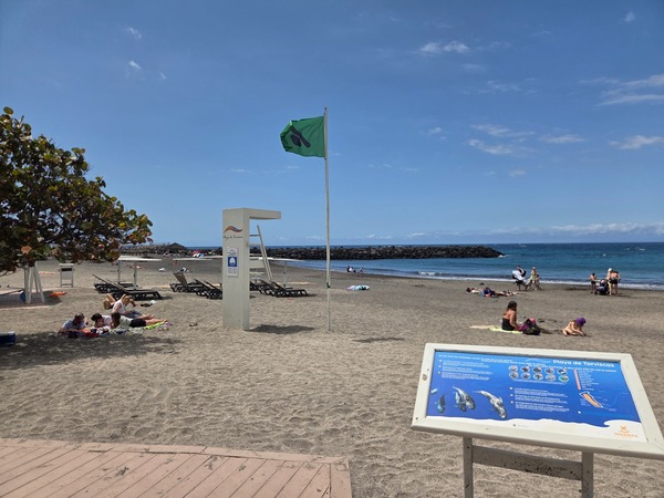 Playa de Torviscas con bandera verde, arena oscura, personas tomando el sol y el mar tranquilo en la costa de Tenerife.