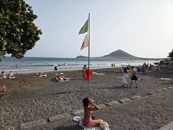 Playa de El Médano con arena oscura, bañistas y la montaña de Montaña Roja al fondo.