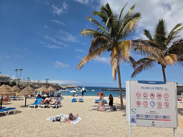 Vista de Playa La Pinta con arena clara, tumbonas, palmeras y un cartel informativo frente al mar.