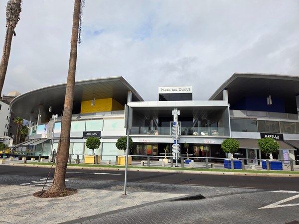 Fachada de Plaza del Duque en Costa Adeje con diseño moderno, escaparates y palmeras frente al edificio.