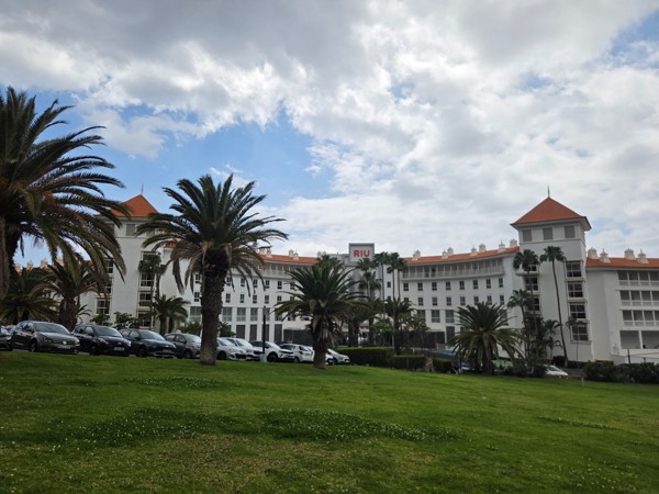 Vista del hotel Riu con fachada blanca, tejados rojizos, jardines y varias palmeras delante del edificio.