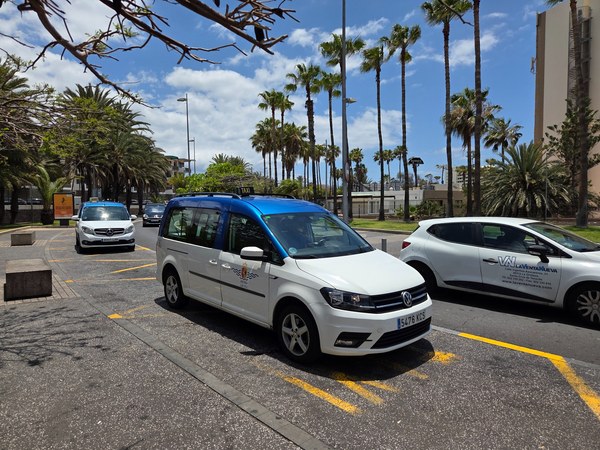 Taxi blanco y azul de Arona estacionado en una avenida con palmeras y otros vehículos en Costa Adeje.